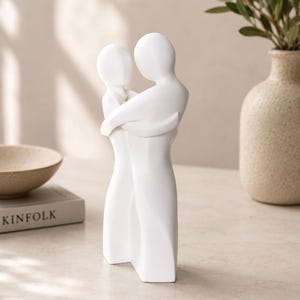 May include: A white ceramic sculpture depicting two figures embracing. The figures are stylized with smooth, rounded forms, conveying a sense of closeness. The sculpture is placed on a light-colored surface, with a neutral background.