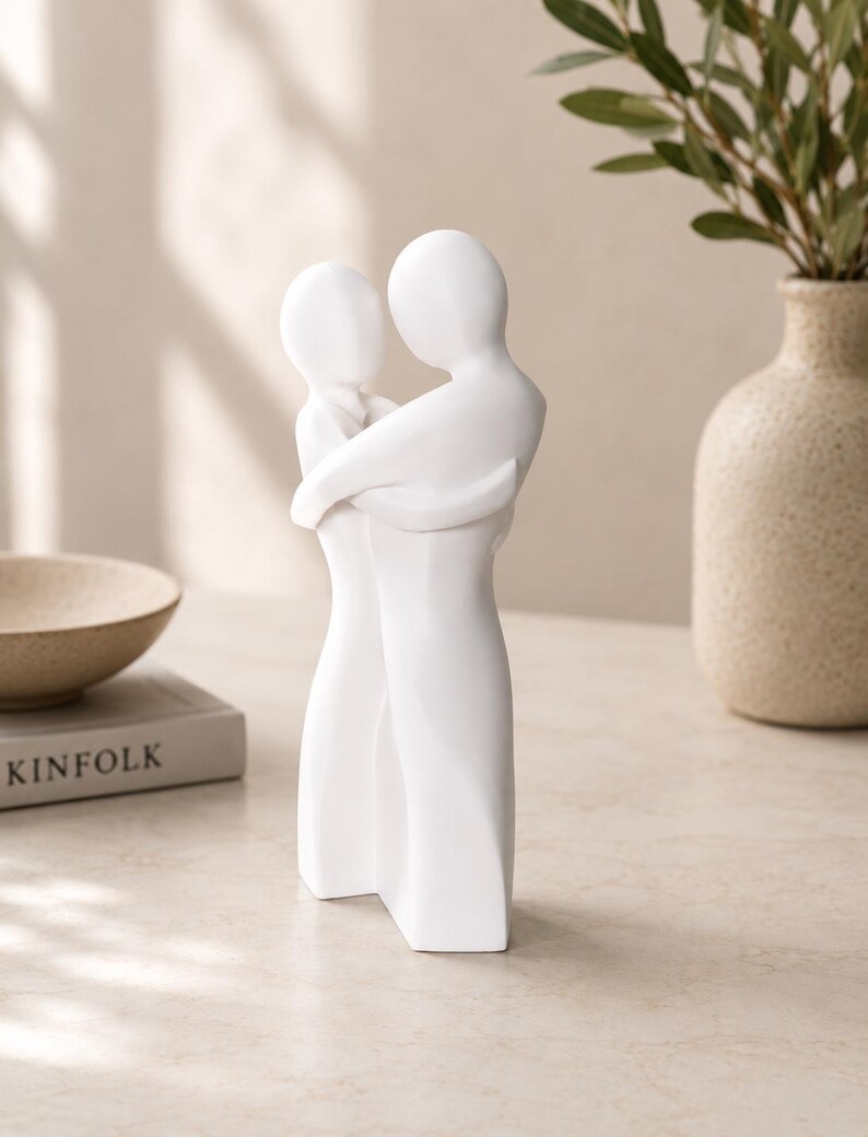 May include: A white ceramic sculpture depicting two figures embracing. The figures are stylized with smooth, rounded forms, conveying a sense of closeness. The sculpture is placed on a light-colored surface, with a neutral background.
