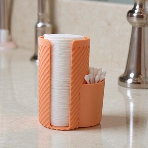 May include: An orange plastic bathroom organizer holds a stack of white cotton pads and a small compartment with cotton swabs. The organizer has a ribbed design and sits on a light-colored countertop.
