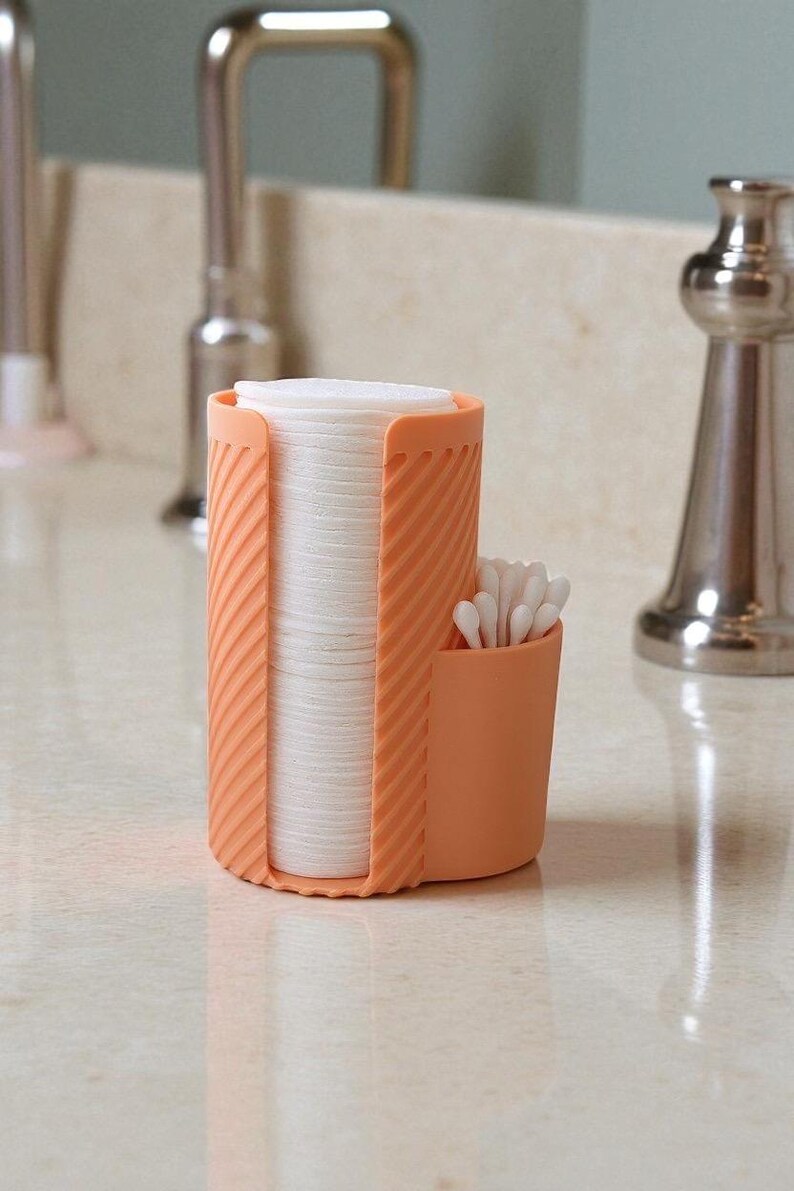 May include: An orange plastic bathroom organizer holds a stack of white cotton pads and a small compartment with cotton swabs. The organizer has a ribbed design and sits on a light-colored countertop.
