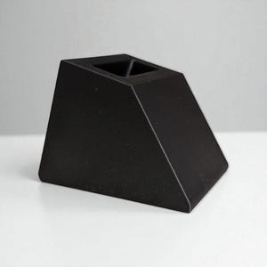 以下が含まれることがあります： 上部に正方形の開口部がある黒色の幾何学的なブロック。オブジェクトは角度の付いた側面を持ち、固体でマットな素材で作られています。白い表面に配置されています。デザインはミニマリストでモダンです。