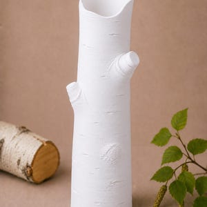 Może przedstawiać: Biały ceramiczny wazon w kształcie pnia brzozy, z fakturą kory i małymi kikutami gałęzi. Wazon ma nieregularny g&oacute;rny otw&oacute;r. Fragment pnia brzozy znajduje się w tle.