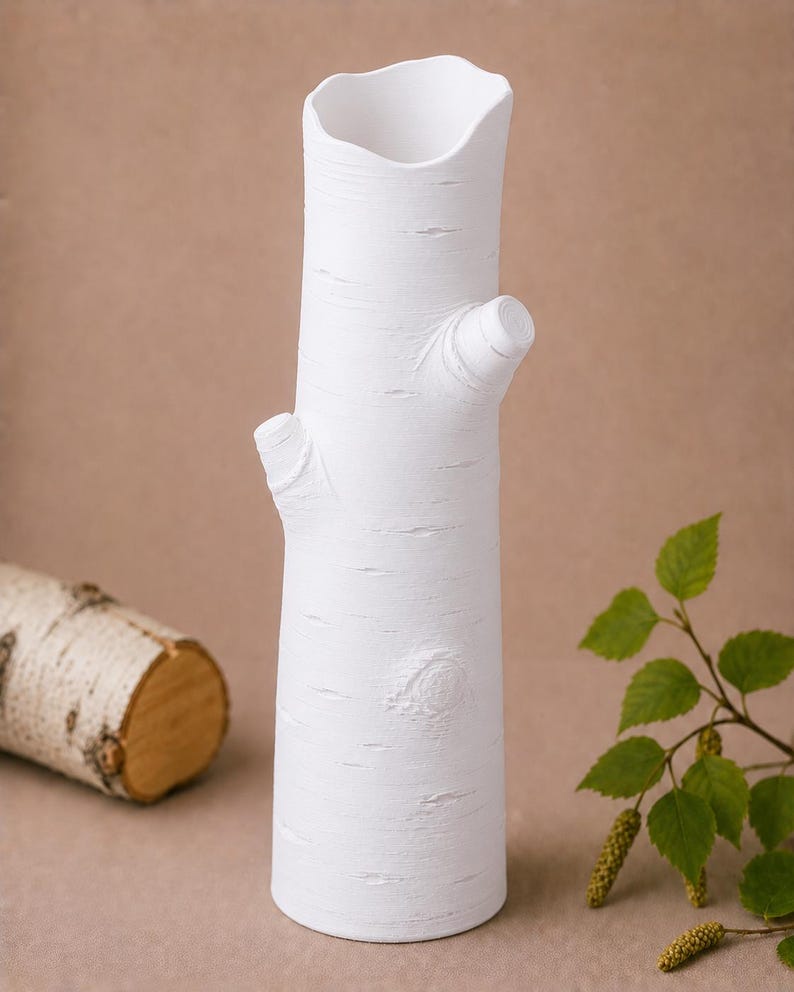 Może przedstawiać: Biały ceramiczny wazon w kształcie pnia brzozy, z fakturą kory i małymi kikutami gałęzi. Wazon ma nieregularny g&oacute;rny otw&oacute;r. Fragment pnia brzozy znajduje się w tle.