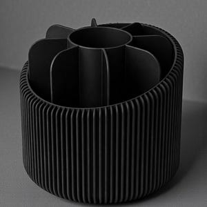 May include: A black, cylindrical desk organizer with a ribbed exterior and interior compartments. The organizer has a central circular section and several curved dividers, creating multiple storage spaces. The design is modern and minimalist.