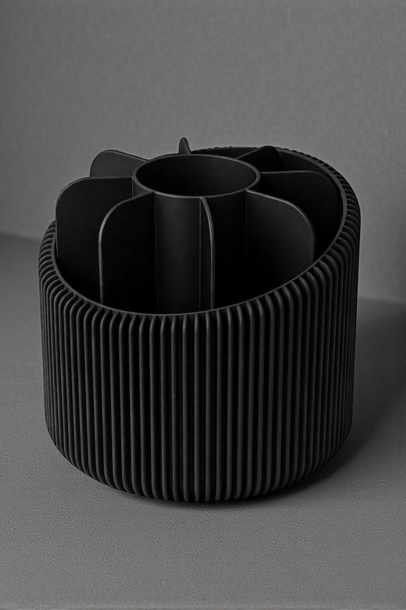 May include: A black, cylindrical desk organizer with a ribbed exterior and interior compartments. The organizer has a central circular section and several curved dividers, creating multiple storage spaces. The design is modern and minimalist.