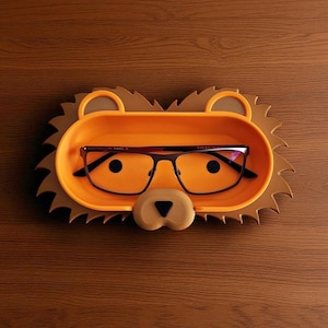 May include: An orange lion-shaped glasses holder with a brown rim and a beige nose. The holder contains a pair of black-framed eyeglasses. The design is on a wooden surface.