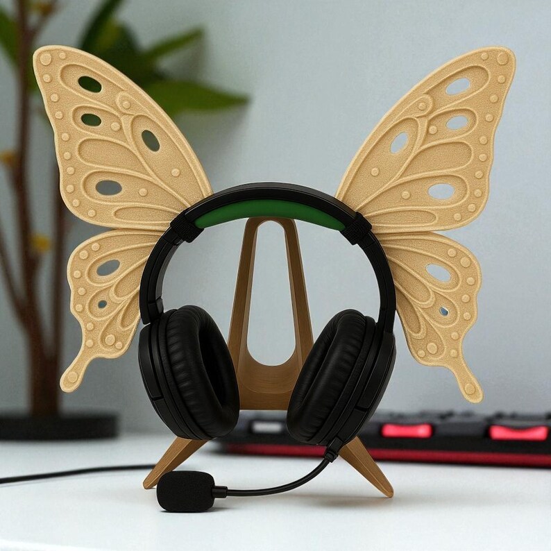 May include: A black headset with a green headband sits on a beige butterfly-shaped headphone stand. The stand has a wooden appearance and is designed to hold the headset. The butterfly wings have a detailed, textured design.