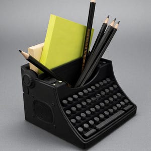 May include: A black desk organizer shaped like a vintage typewriter. The organizer holds several black pencils, a yellow sticky note pad, and a small beige pad. The typewriter has a full keyboard and a speaker-like detail on the side.