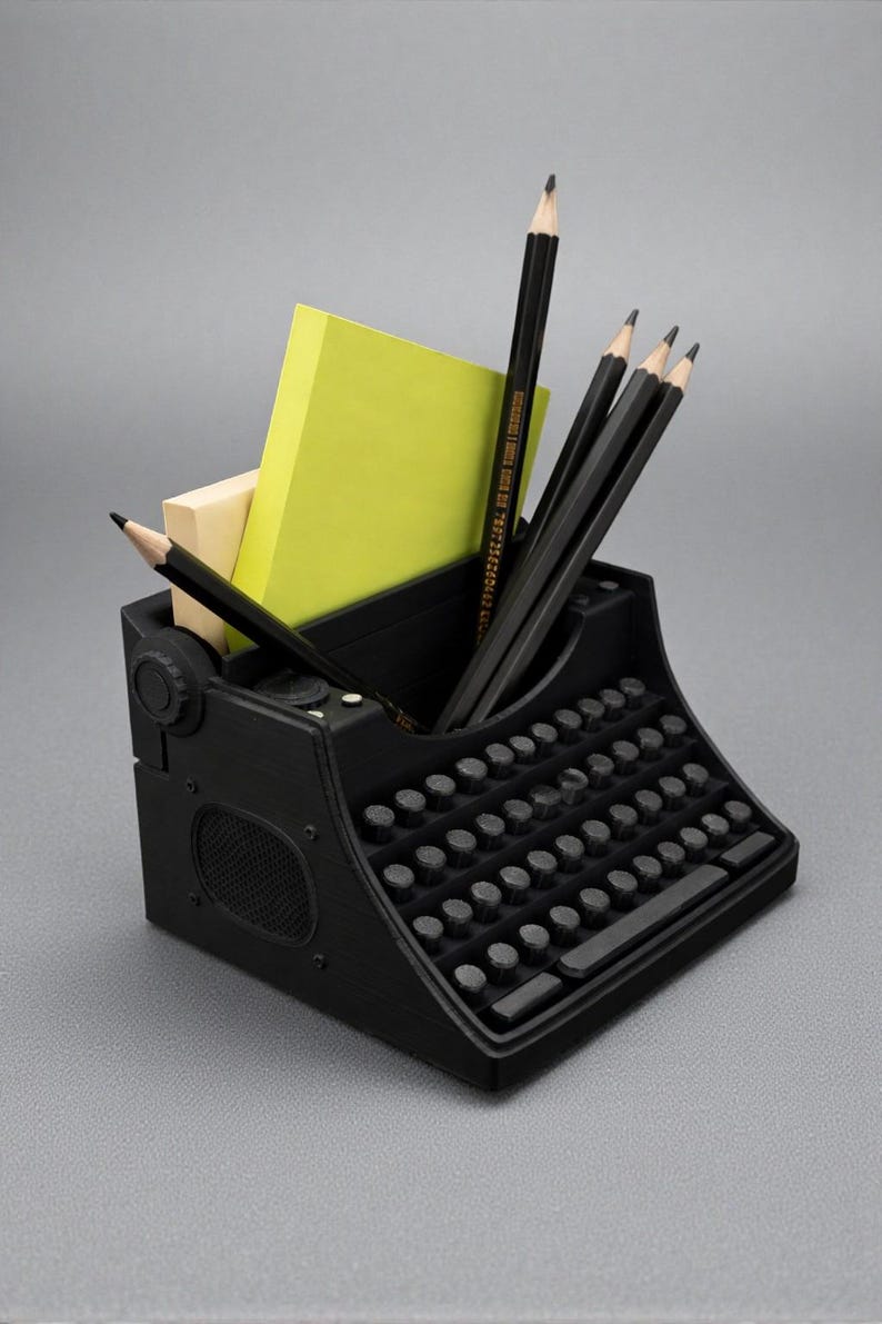 May include: A black desk organizer shaped like a vintage typewriter. The organizer holds several black pencils, a yellow sticky note pad, and a small beige pad. The typewriter has a full keyboard and a speaker-like detail on the side.