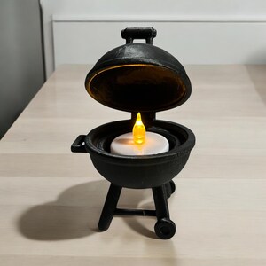 May include: A black miniature barbecue grill with a lid that opens. Inside, a white disc holds a flickering yellow LED candle. The grill has a handle on top and sits on three legs with a wheel on one side. A unique decorative item.