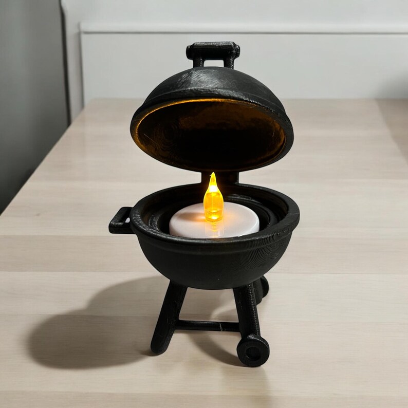 May include: A black miniature barbecue grill with a lid that opens. Inside, a white disc holds a flickering yellow LED candle. The grill has a handle on top and sits on three legs with a wheel on one side. A unique decorative item.