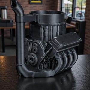 May include: A black, 3D-printed mug shaped like a V8 engine. The mug features a detailed design with exhaust pipes, the number "V8", and other engine components. The mug has a handle and a cylindrical opening for beverages.