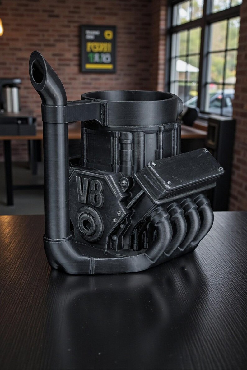May include: A black, 3D-printed mug shaped like a V8 engine. The mug features a detailed design with exhaust pipes, the number "V8", and other engine components. The mug has a handle and a cylindrical opening for beverages.