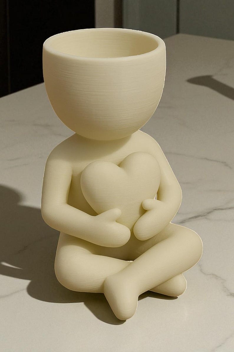May include: A cream-colored figurine of a person seated with legs crossed, holding a heart-shaped object. The figure has a large, open-topped head, resembling a planter. The figurine is made of a smooth, matte material.
