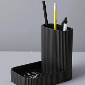 May include: A black desk organizer with a ribbed texture. It features a tall, oval-shaped section for pens and pencils, and a lower tray for paperclips. A yellow pencil, black pen, and black stick are inside.