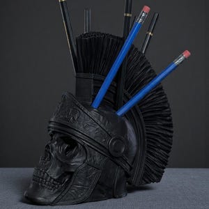 Puede incluir: Un organizador de escritorio negro con forma de calavera con un dise&ntilde;o de casco romano. El organizador contiene l&aacute;pices y bol&iacute;grafos, incluidos l&aacute;pices azules con gomas rosas. El dise&ntilde;o presenta un casco detallado y un acabado mate texturizado.