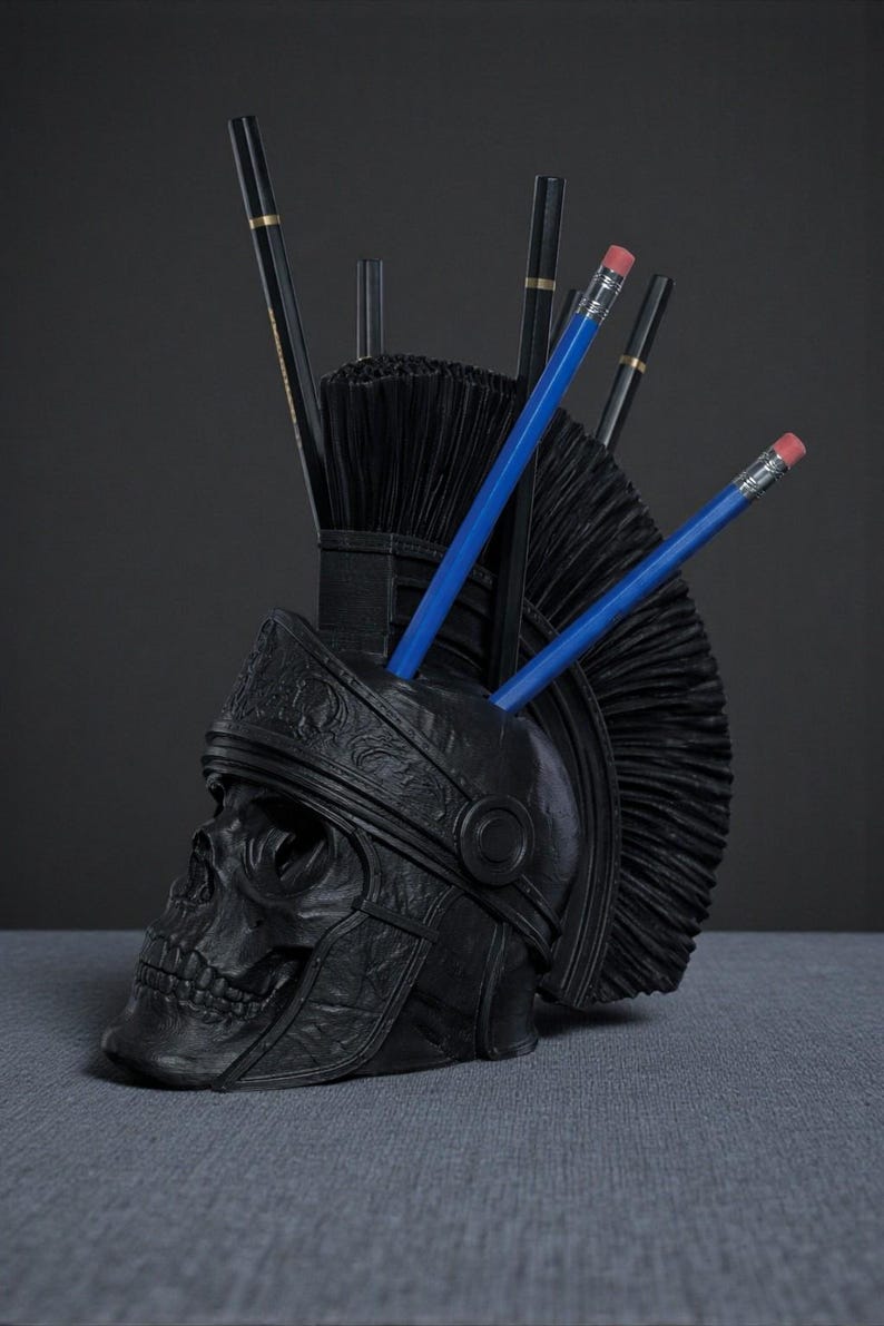 Puede incluir: Un organizador de escritorio negro con forma de calavera con un dise&ntilde;o de casco romano. El organizador contiene l&aacute;pices y bol&iacute;grafos, incluidos l&aacute;pices azules con gomas rosas. El dise&ntilde;o presenta un casco detallado y un acabado mate texturizado.