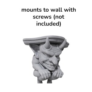 K&ouml;nnte beinhalten: Graue Gargoyle-Regalhalterung mit teuflischem Design. Die Halterung hat H&ouml;rner und ein grimmiges Gesicht und ist f&uuml;r die Wandmontage mit Schrauben vorgesehen. Der Text "mounts to wall with screws (not included)" befindet sich &uuml;ber dem Gargoyle.