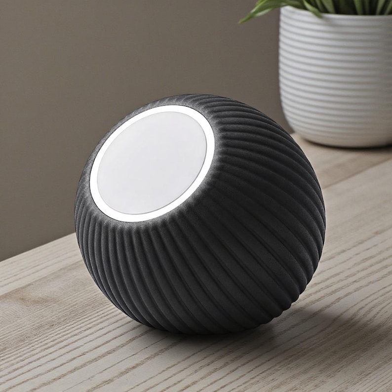 May include: A dark gray, spherical lamp with a textured, ribbed surface. The lamp has a bright, circular light on top. The lamp is sitting on a light-colored wooden surface. A white potted plant is in the background.