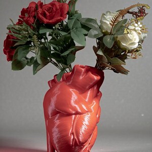 May include: A glossy, red heart-shaped vase holding bouquets of red roses and white roses with green foliage. The vase has a detailed anatomical design, and the flowers are arranged to create a striking visual contrast.