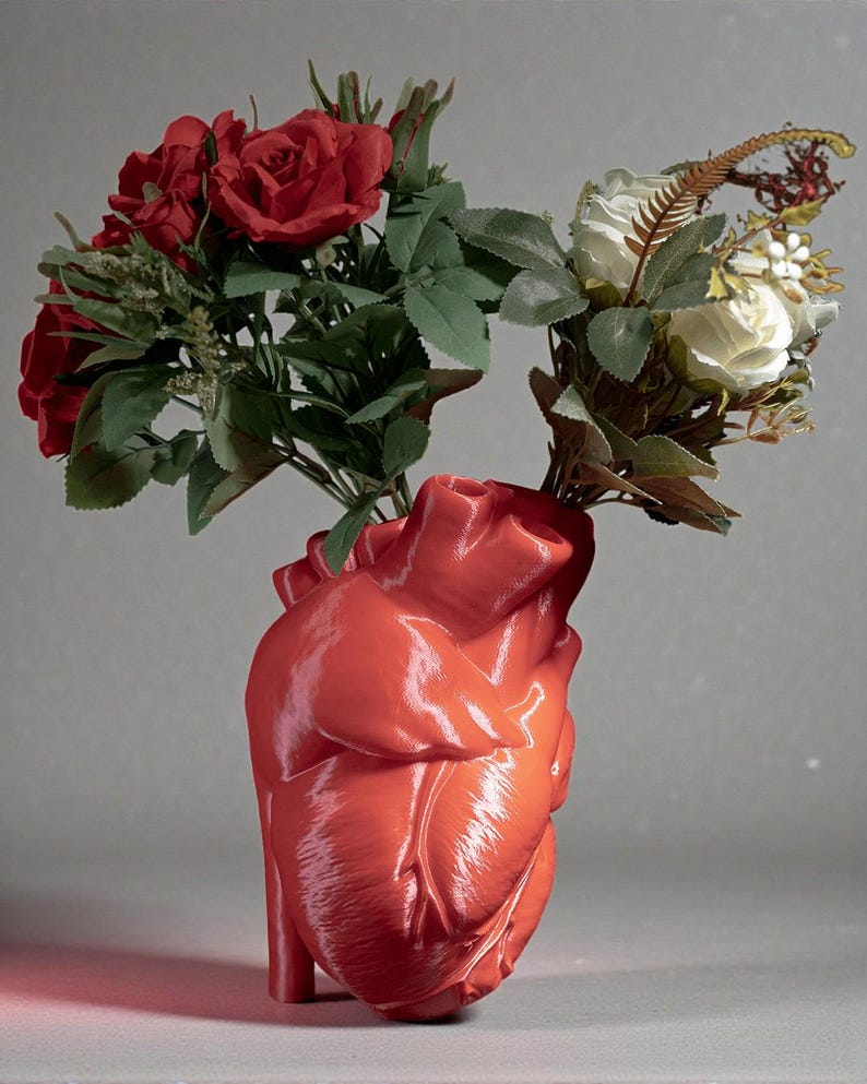 May include: A glossy, red heart-shaped vase holding bouquets of red roses and white roses with green foliage. The vase has a detailed anatomical design, and the flowers are arranged to create a striking visual contrast.