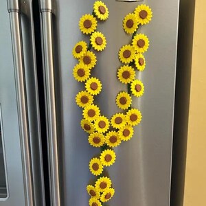 May include: A collection of yellow sunflower magnets with brown centers arranged in a vertical pattern on a stainless steel refrigerator. The magnets are clustered together, creating a decorative floral display.