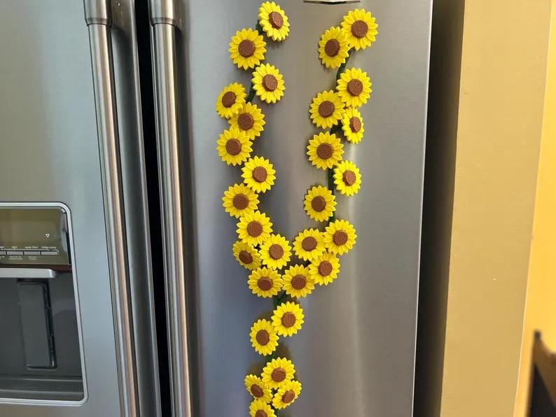 May include: A collection of yellow sunflower magnets with brown centers arranged in a vertical pattern on a stainless steel refrigerator. The magnets are clustered together, creating a decorative floral display.