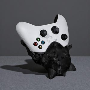 May include: A white Xbox controller sits on a black, spiky, 3D-printed stand. The controller has coloured buttons and black control sticks. The stand is in the shape of a creature. The background is a solid grey.