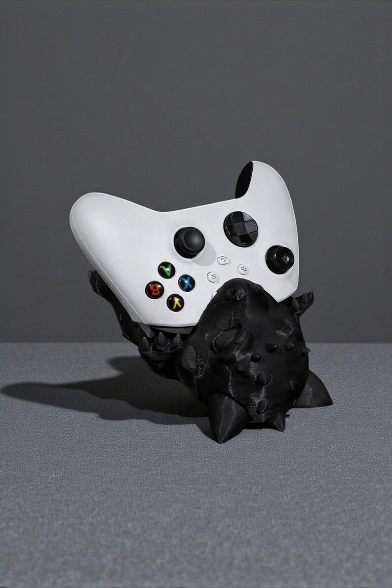 May include: A white Xbox controller sits on a black, spiky, 3D-printed stand. The controller has coloured buttons and black control sticks. The stand is in the shape of a creature. The background is a solid grey.