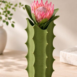 May include: A green cactus-shaped vase holds a large pink protea flower with green leaves. The vase has a textured surface and is decorated with small, yellow-green protrusions. The flower is in full bloom.