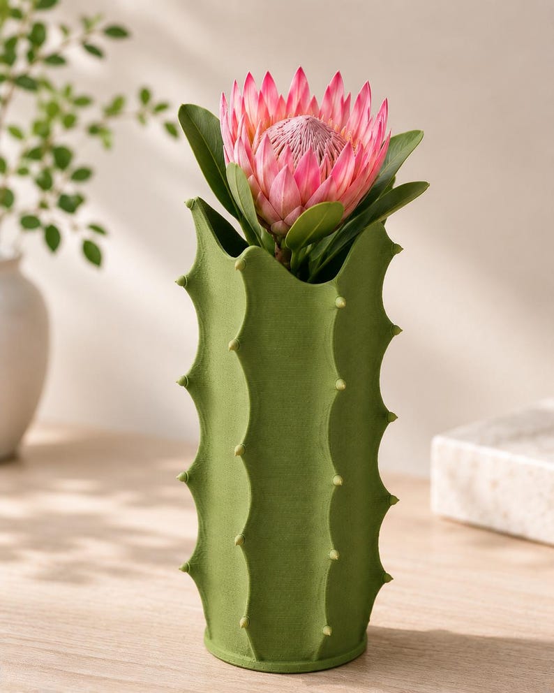 May include: A green cactus-shaped vase holds a large pink protea flower with green leaves. The vase has a textured surface and is decorated with small, yellow-green protrusions. The flower is in full bloom.
