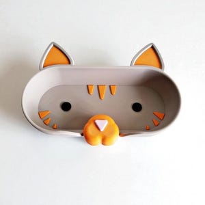 May include: A grey cat-shaped wall shelf with orange and black detailing. The shelf is designed as a cat's face, with ears, eyes, nose, and whiskers. The shelf is a rounded rectangle shape and is designed to hold small items.