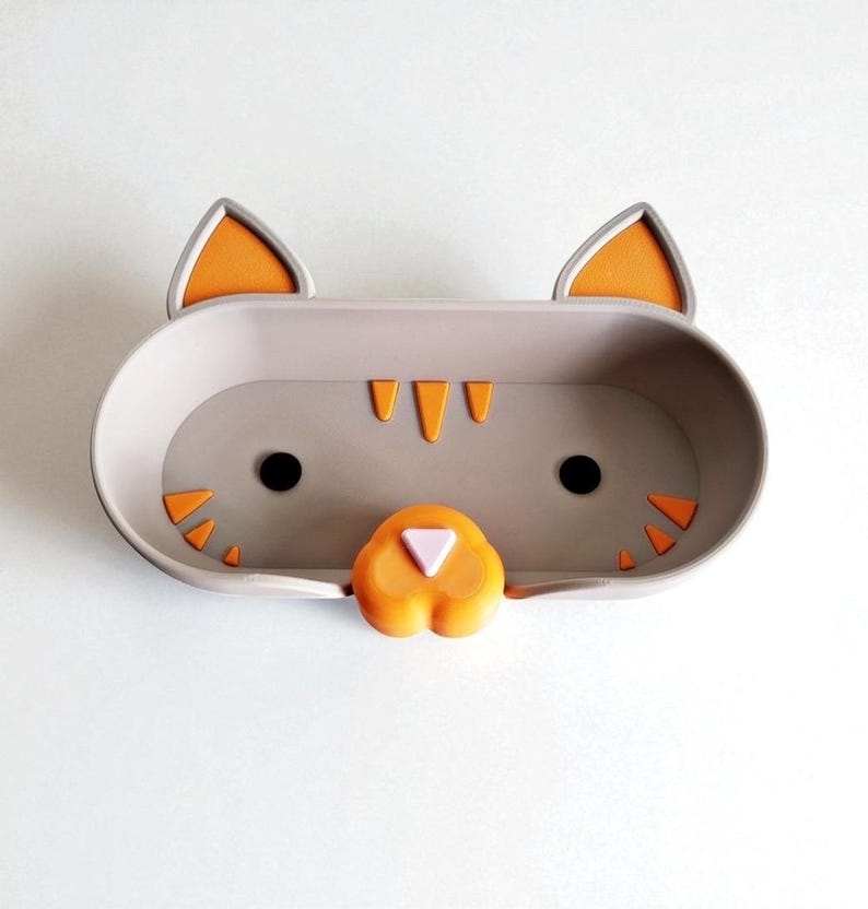 May include: A grey cat-shaped wall shelf with orange and black detailing. The shelf is designed as a cat's face, with ears, eyes, nose, and whiskers. The shelf is a rounded rectangle shape and is designed to hold small items.