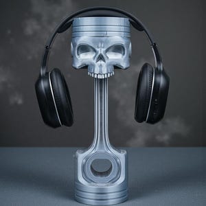 May include: A silver-colored headphone stand shaped like a piston with a skull design. The stand holds a pair of black over-ear headphones. The background is a gradient of gray.