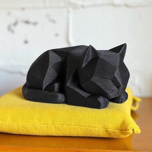 May include: A black geometric cat sculpture resting on a bright yellow square pillow. The cat is in a sleeping position, with its ears perked up. The pillow is made of a textured fabric. The background is a white brick wall.