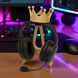 May include: A black headset with a microphone rests on a wooden stand, topped with a gold-colored crown. The stand is on a wooden surface, with a keyboard and monitor in the background.