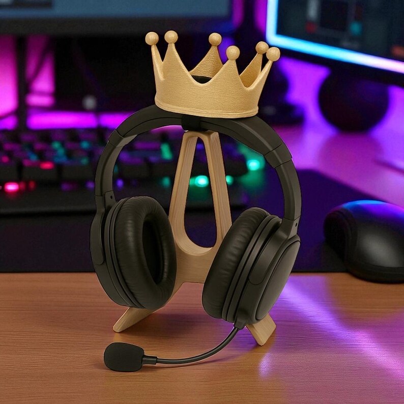 May include: A black headset with a microphone rests on a wooden stand, topped with a gold-colored crown. The stand is on a wooden surface, with a keyboard and monitor in the background.