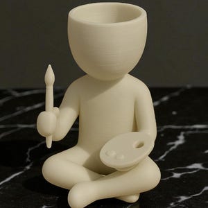 May include: A cream-colored figurine of a seated artist holding a paintbrush and palette. The figure has a large, open head and is in a meditative pose. The artist's palette has two paint wells. The figurine is on a black and white marble surface.