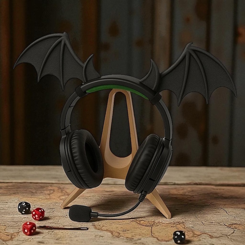 May include: Black gaming headphones with a bat wing and horn design, resting on a wooden stand. The headphones have a built-in microphone. Three dice are on the table, two red and one black.