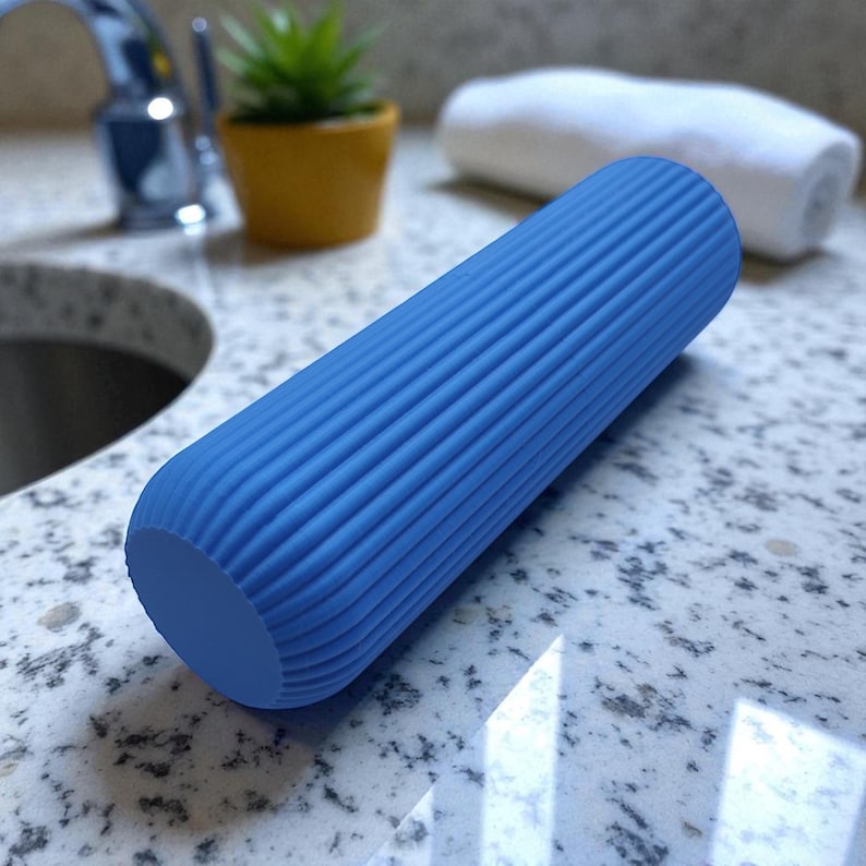 Pode incluir: Um rolo de espuma azul com uma superf&iacute;cie canelada. O rolo cil&iacute;ndrico &eacute; projetado para massagem muscular e est&aacute; sobre uma bancada salpicada. Uma pequena planta em vaso e uma toalha branca est&atilde;o no fundo.