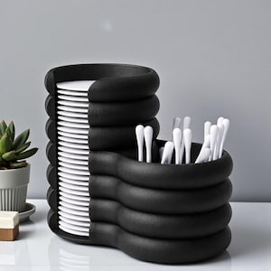 Pode incluir: Um porta-pratos e talheres preto e moderno. O porta-pratos tem um design empilhado e ondulado, segurando uma pilha de pratos brancos. O porta-talheres tem um design semelhante e cont&eacute;m talheres brancos. Uma planta suculenta num vaso cinzento est&aacute; ao fundo.