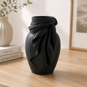 May include: A matte black decorative vase with a draped fabric design around the neck. The vase has a smooth, rounded shape and sits on a wooden table. A white vase and books are in the background.