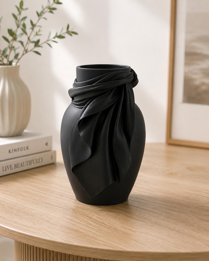 May include: A matte black decorative vase with a draped fabric design around the neck. The vase has a smooth, rounded shape and sits on a wooden table. A white vase and books are in the background.