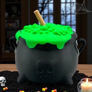 May include: A black cauldron with a green, bubbling, liquid-like substance on top and a wooden stirring stick. The cauldron has a skull design on the front and handles on the sides. Candy corn and a lit candle are in the foreground.