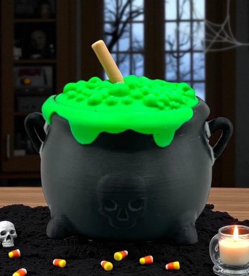 May include: A black cauldron with a green, bubbling, liquid-like substance on top and a wooden stirring stick. The cauldron has a skull design on the front and handles on the sides. Candy corn and a lit candle are in the foreground.