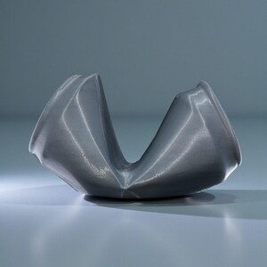 May include: A gray, abstract sculpture with a curved, open design. The object has a textured surface and appears to be made of a metallic material. The sculpture is set against a light gray background, with soft lighting.