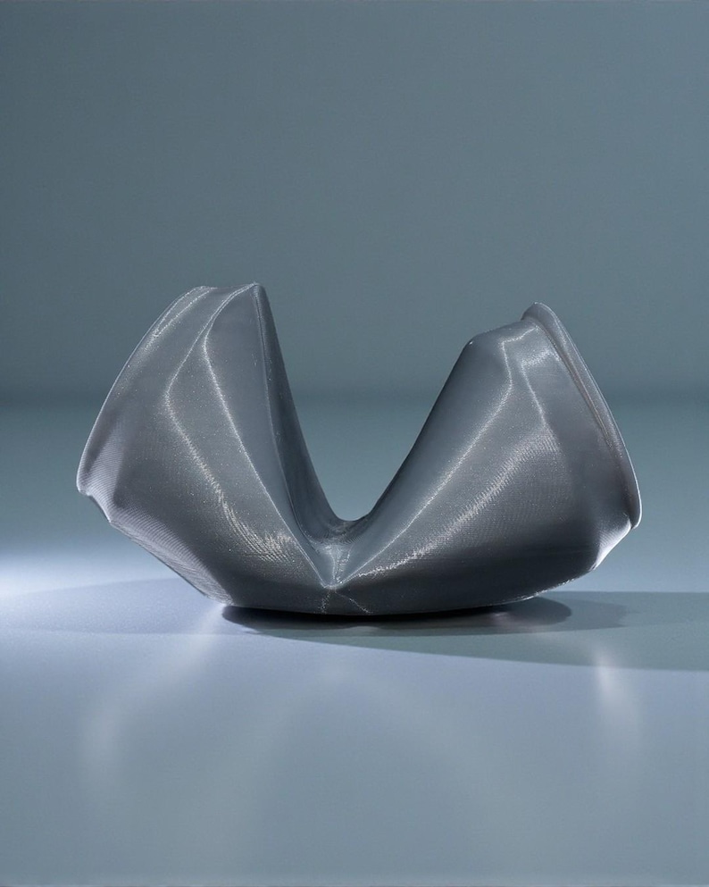 May include: A gray, abstract sculpture with a curved, open design. The object has a textured surface and appears to be made of a metallic material. The sculpture is set against a light gray background, with soft lighting.