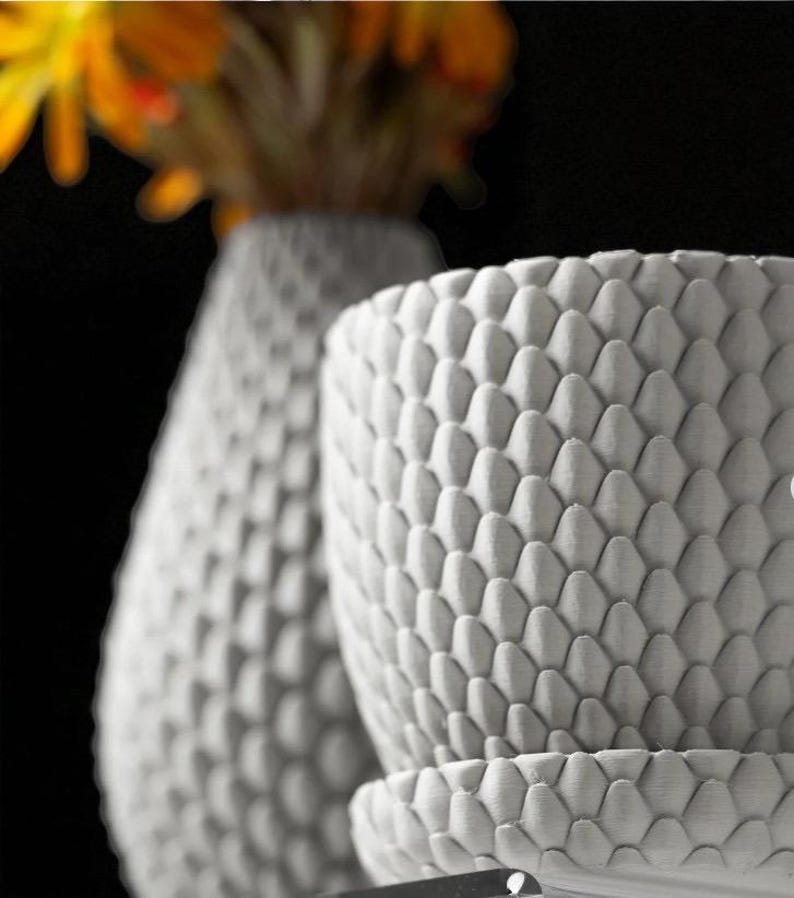 Pu&ograve; includere: Primo piano di due vasi in ceramica bianca strutturata. Il vaso rotondo pi&ugrave; grande ha un piattino abbinato. La superficie di entrambi i vasi &egrave; ricoperta da un motivo ripetitivo a forma di scaglia. Un vaso e dei fiori sfocati sono sullo sfondo.