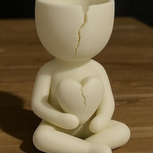 May include: A cream-coloured figurine of a person seated with legs crossed, holding a heart. Both the head and the heart have a crack running through them. The figurine is made of a smooth, matte material.