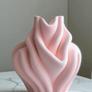 May include: A light pink ceramic vase with a unique, flowing design. The vase has a wide, rounded base that tapers upwards, with the top opening in a wavy, petal-like shape. The smooth, matte finish adds to its modern aesthetic.
